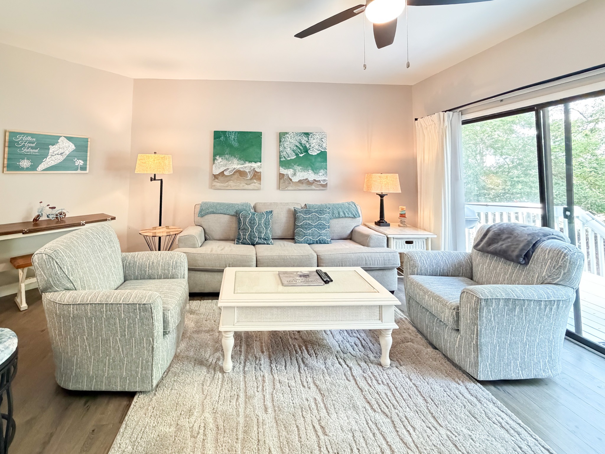 Bright living room with a gray sectional, teal pillows, and a white coffee table under a ceiling fan with coastal decor.