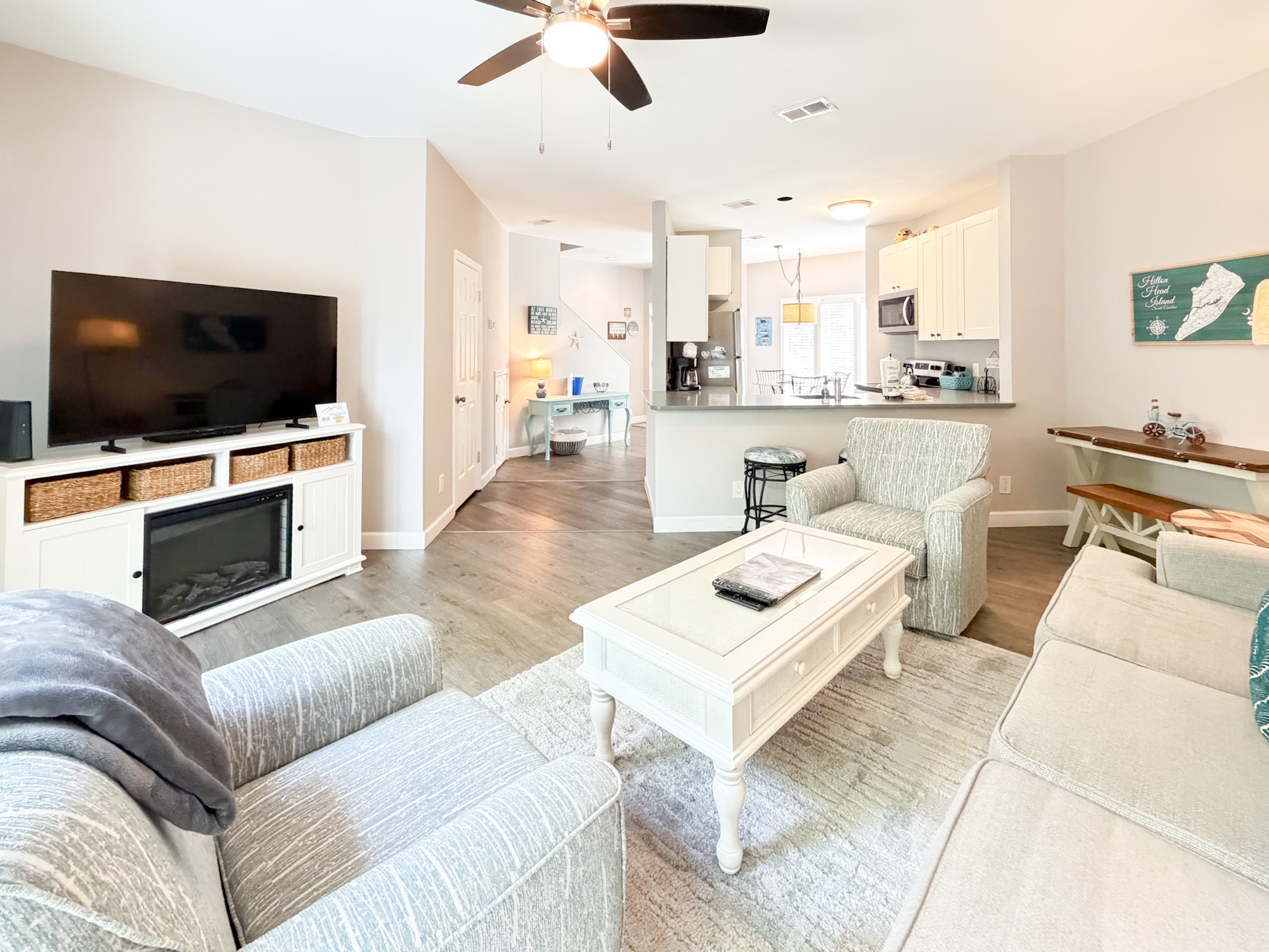 Open living area with a gray sectional, white coffee table, and a wall-mounted TV; kitchen in the background.