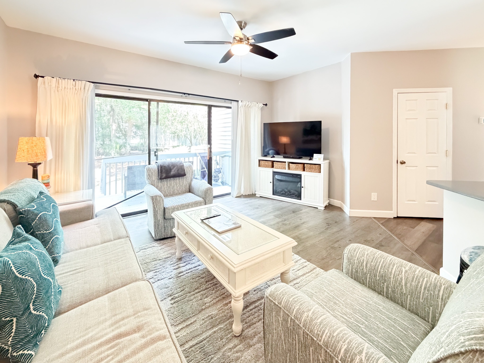 Bright living room with beige walls, sliding glass doors to a patio, white curtains, and a white TV console with a television on top.